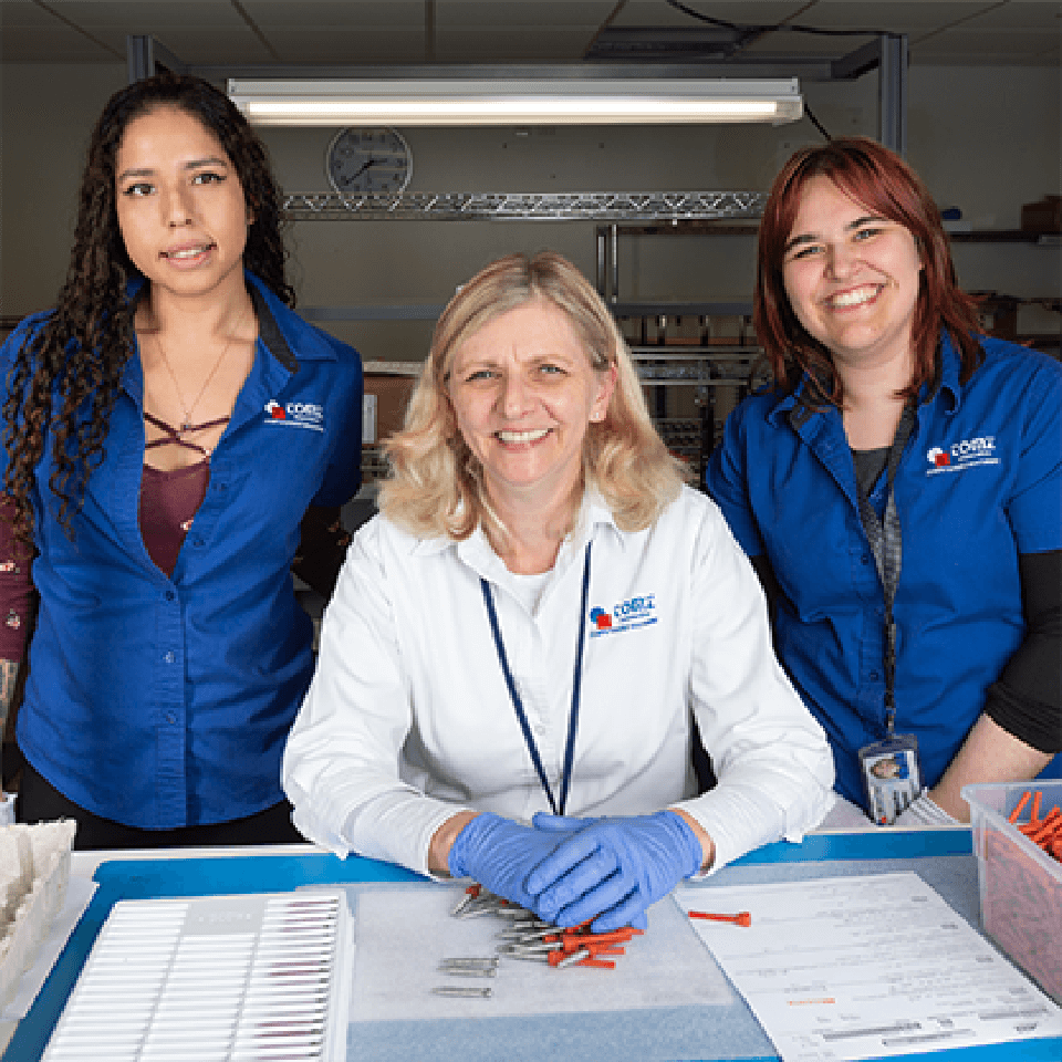 Three women employees, two wearing blue shirts and one wearing a white shirt and blue latex gloves, stand at a table as they smile and perform quality control inspections on specialty screws and other manufactured parts.