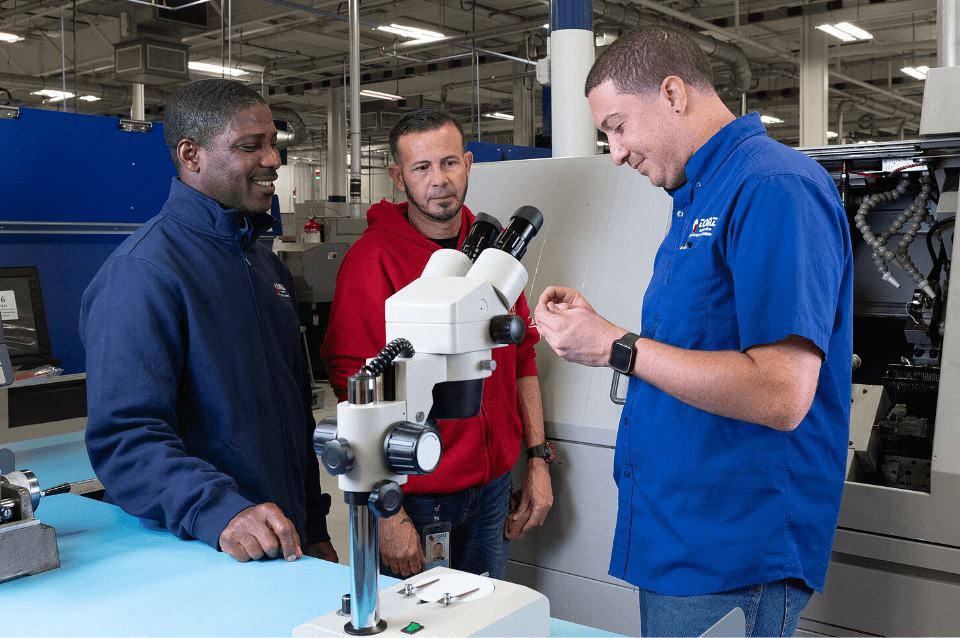 Three employees, two wearing blue shirts and one wearing a red shirt, stand next to a microscope in the machine shop as they look closely at a manufactured part.