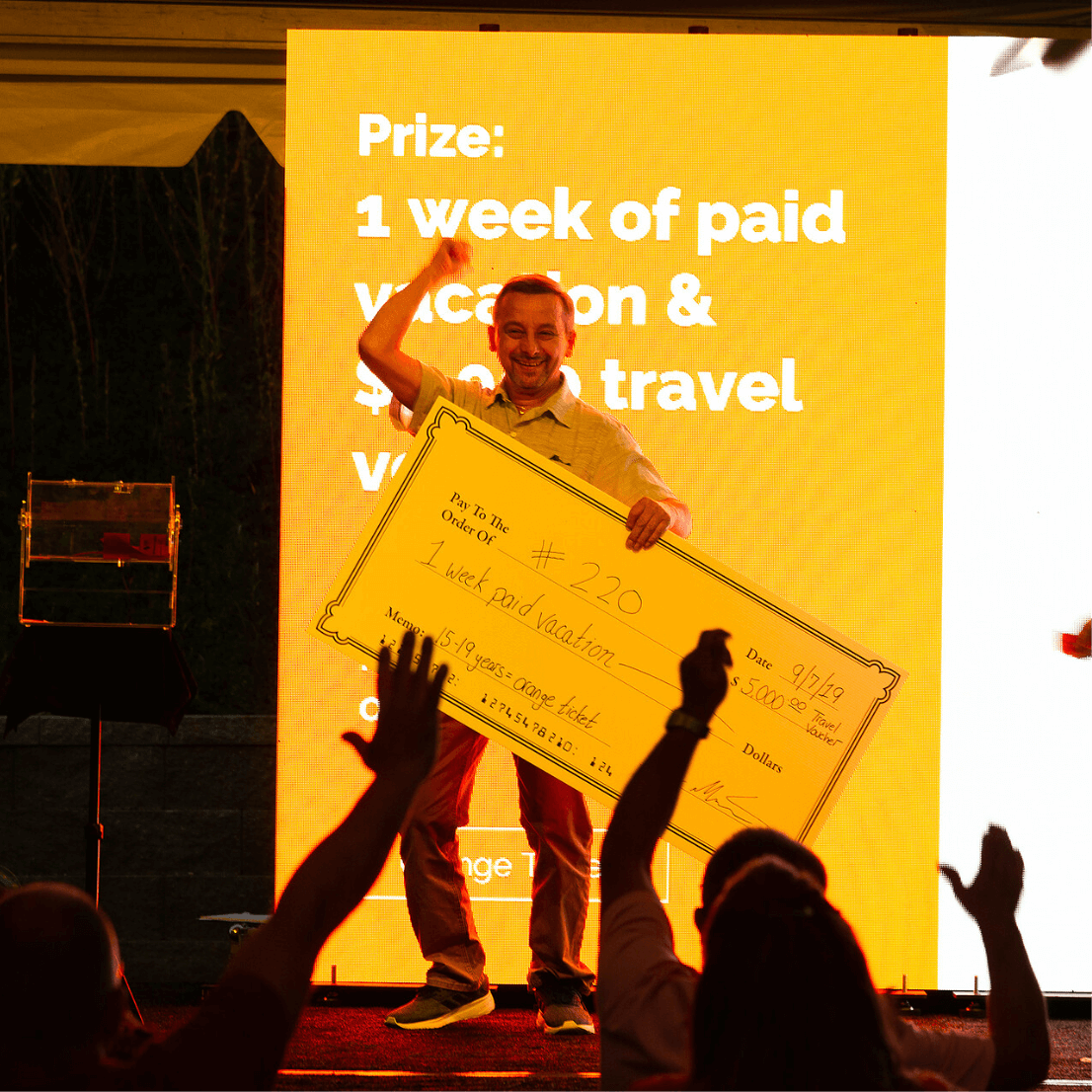 An employee stands in front of a yellow background that says he's won the prize of a week's paid vacation and a $5,000 travel voucher as he holds onto an oversized check stating the same. Other employees in the audience cheer him on.