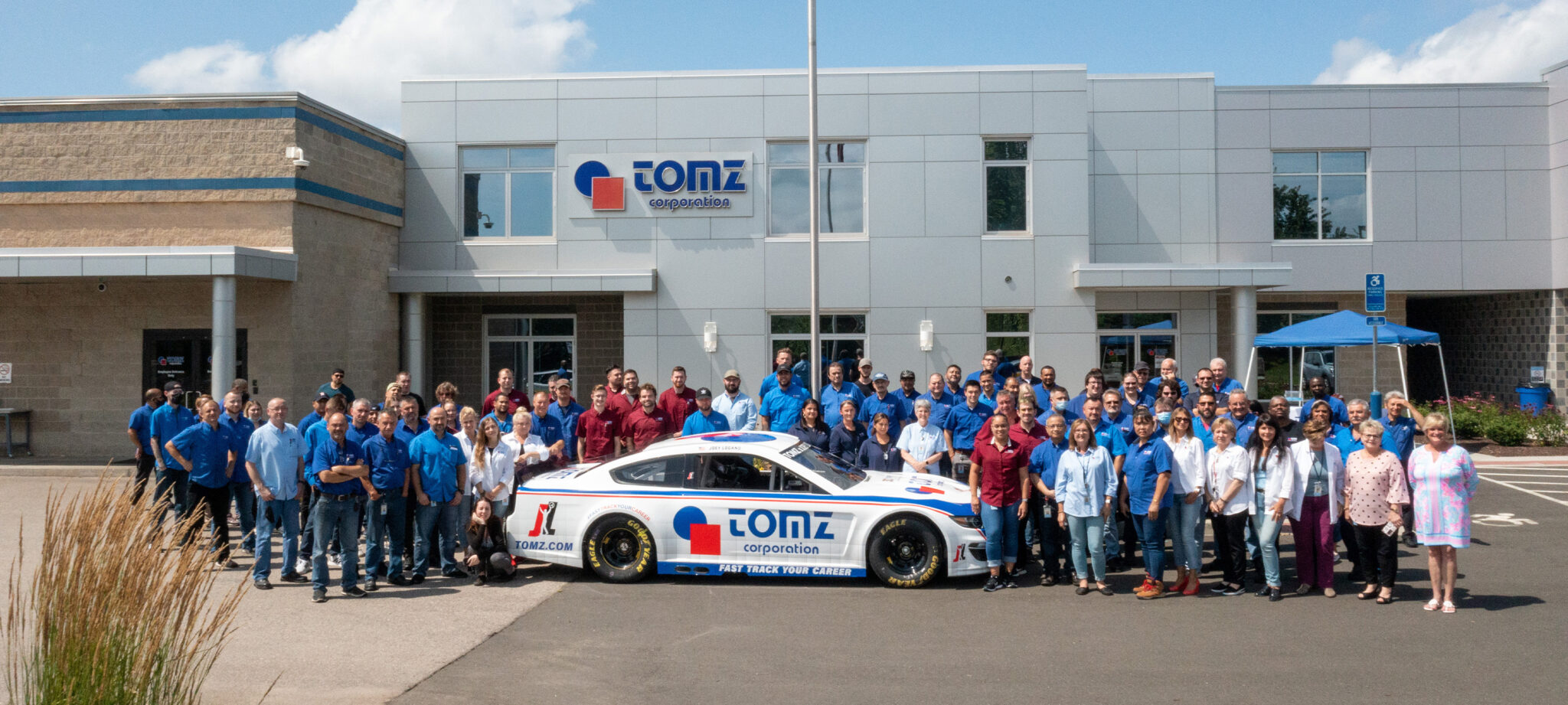 Group photo of the TOMZ Corporation team standing next to a white sports car with the blue and red TOMZ Corporation logo on it in front of the company's headquarters in Berlin, Connecticut.
