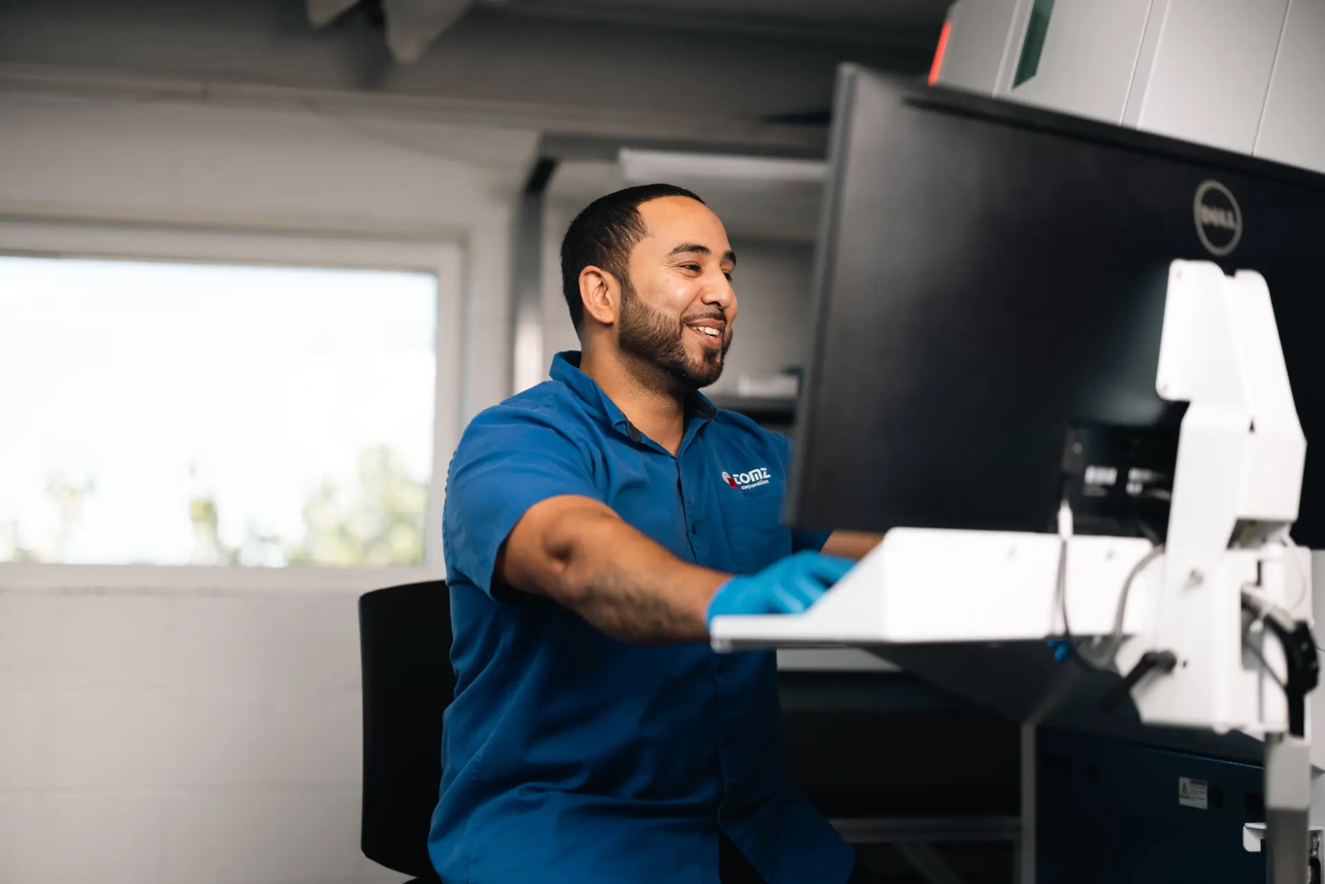 TOMZ Corporation employee wearing a blue uniform and blue latex gloves sits at a desktop computer.